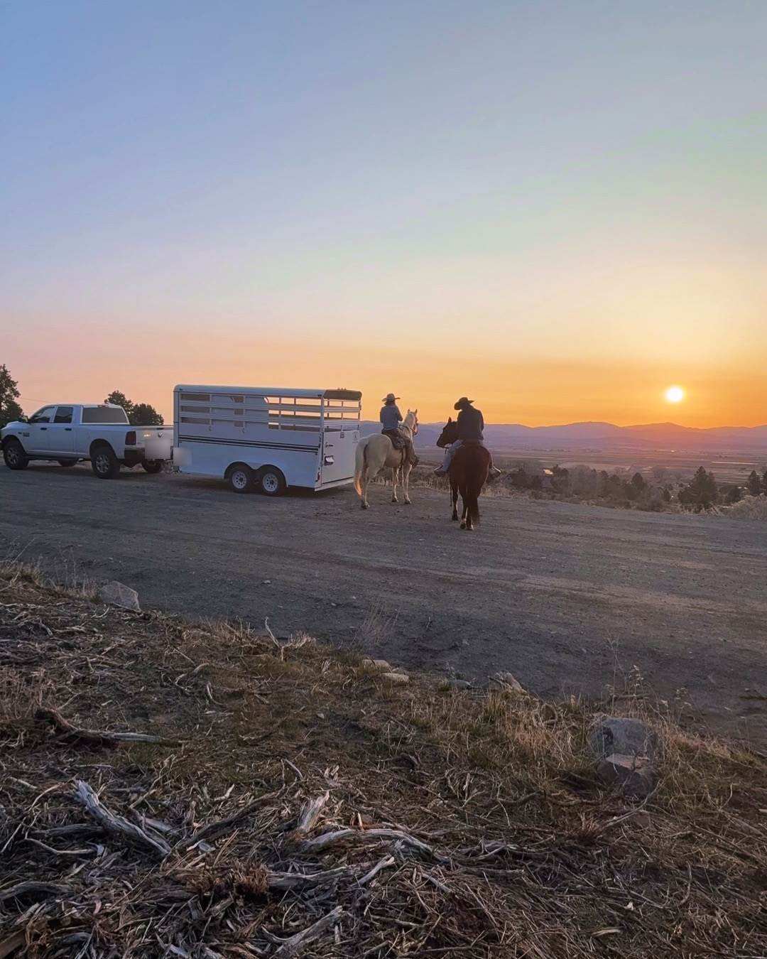 A truck and stock trailer parked at a gravel trailhead as two riders head out at sunset