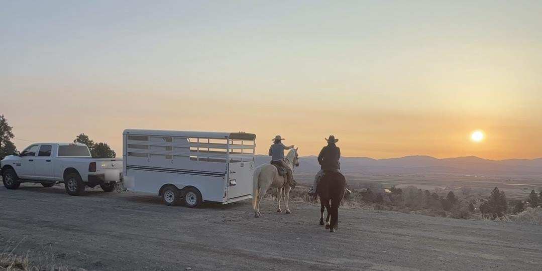 A truck and stock trailer parked at a gravel trailhead as two riders head out toward the sunset